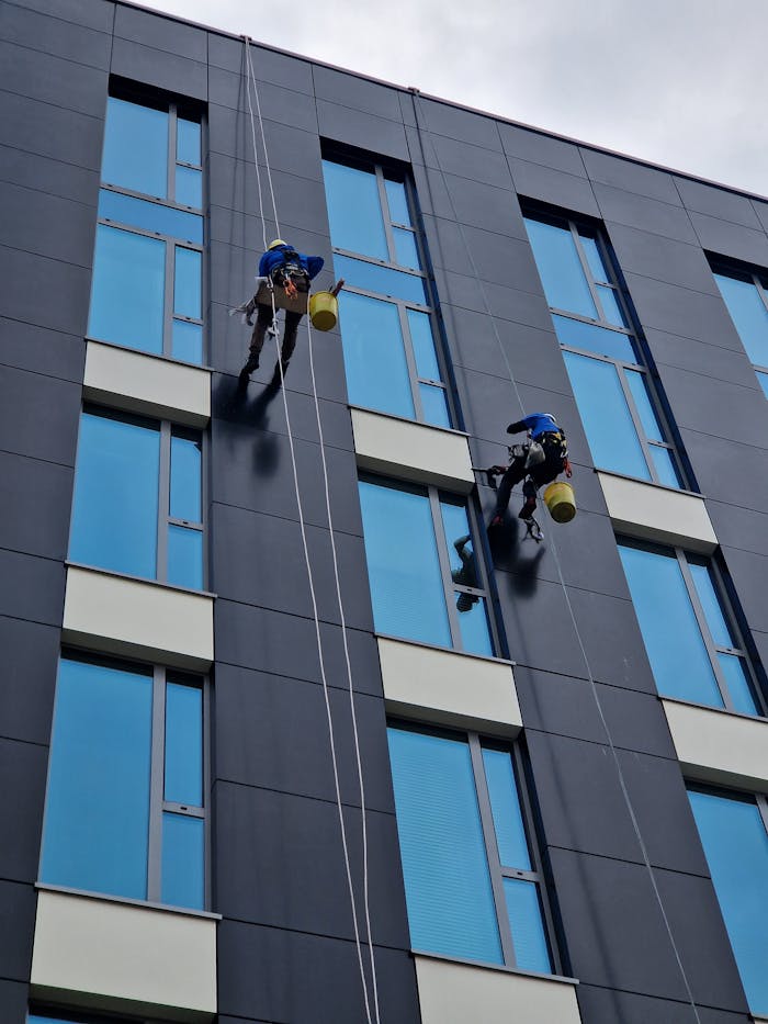 Home Two workers clean a modern building facade at height with safety ropes.