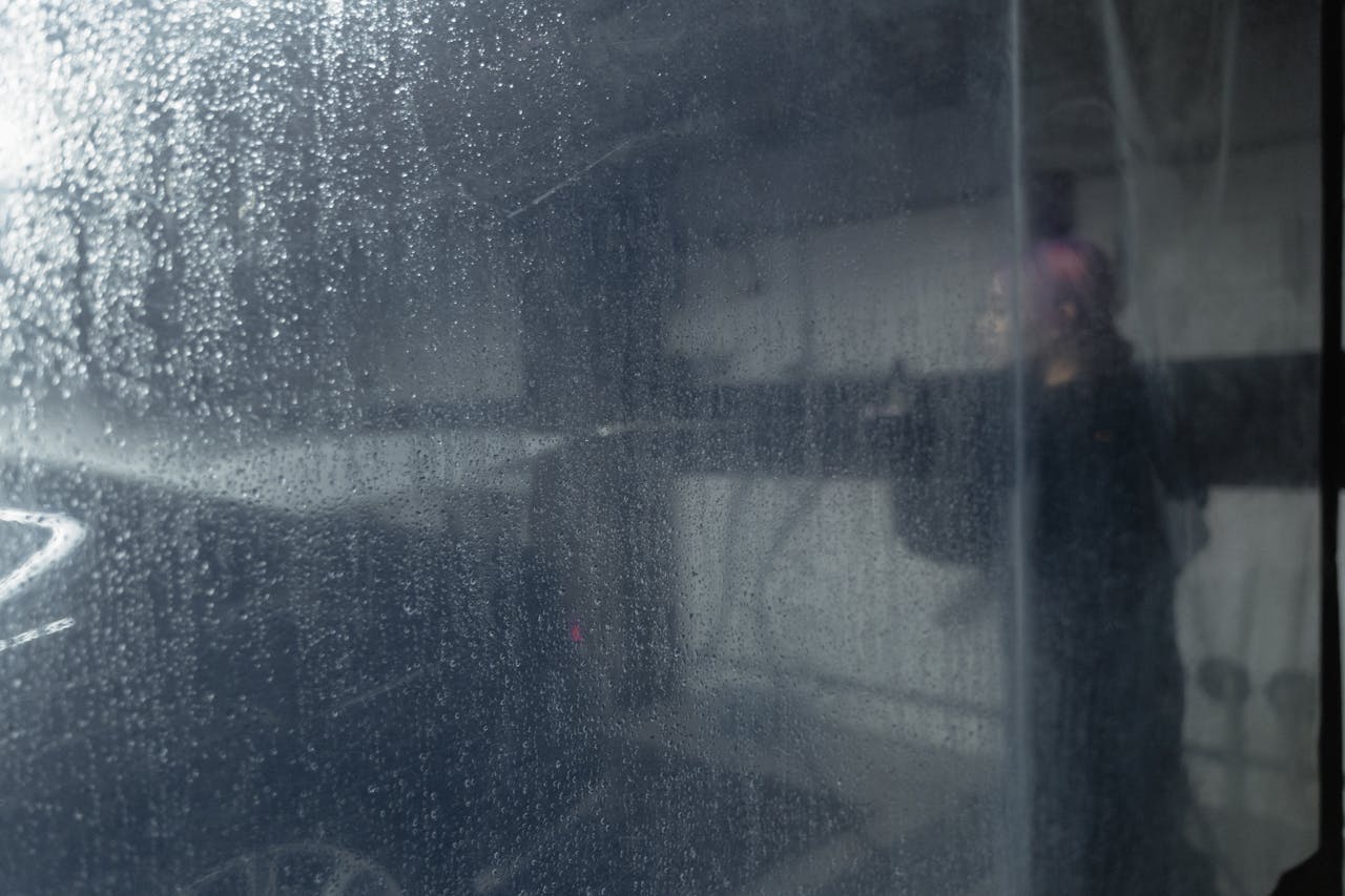 A person washes a car behind a water-streaked plastic curtain, creating a blurred effect.